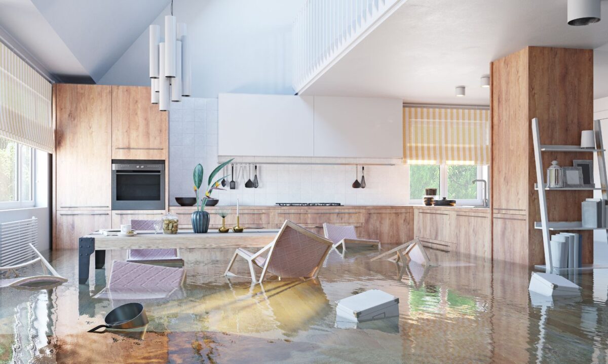 A modern kitchen and dining area with wooden cabinets and furniture partially submerged in water. Chairs are overturned, and various household items float on the water's surface, reflecting the light coming through the windows.