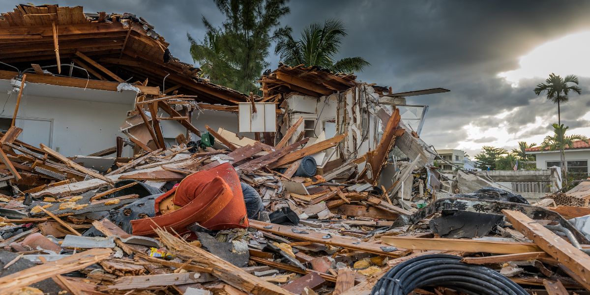 The remains of a destroyed house with debris scattered across the ground. Broken walls, furniture, and roofing material lie in disarray under a cloudy sky, with a few palm trees visible in the background, conveying the aftermath of a severe hurricane.